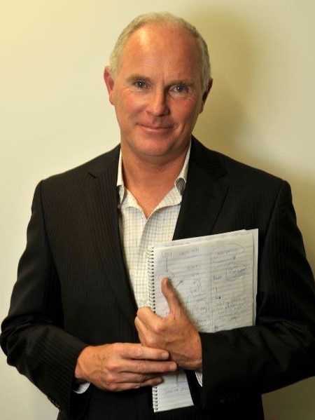 Michael Gordon stands in front of a beige background, smiling and holding a notebook.