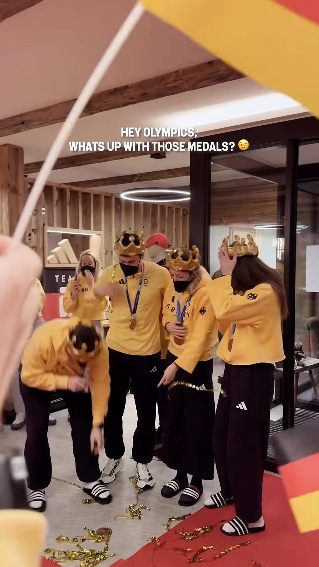 Justus Strelow se abaixa para pegar sua medalha quebrada enquanto os companheiros alemães de biatlo riem.