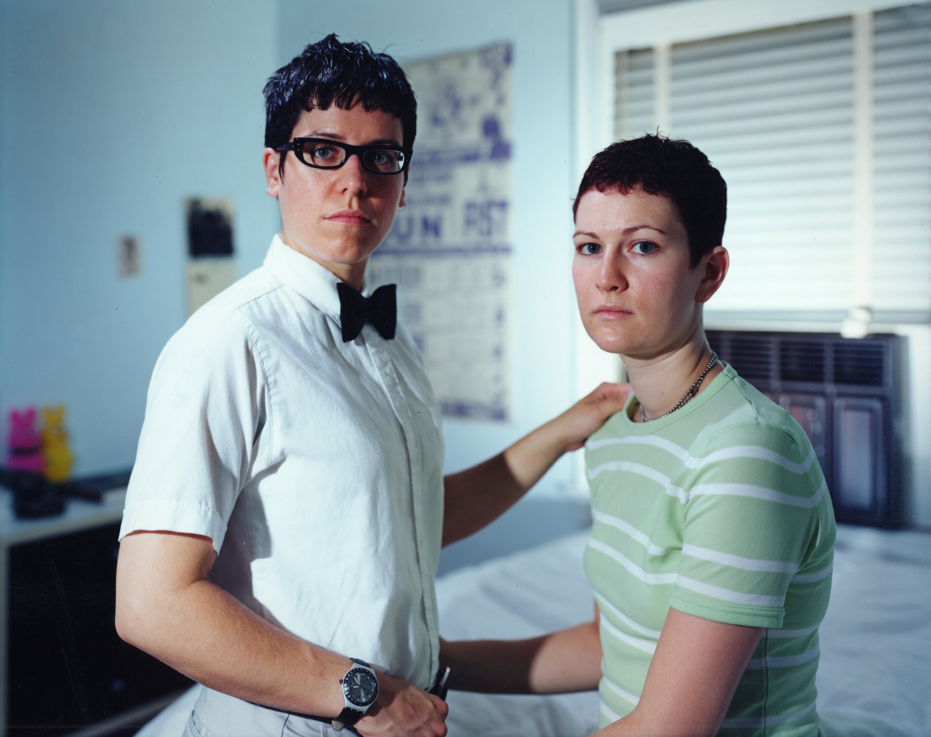 A young lesbian couple stand facing each other, hands on each other's hip and shoulder, heads turned towards the camera.
