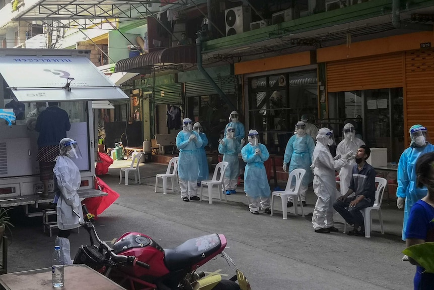 Le persone che indossano dispositivi di protezione individuale completi stanno per strada mentre un uomo seduto su una sedia di plastica viene testato per il coronavirus.
