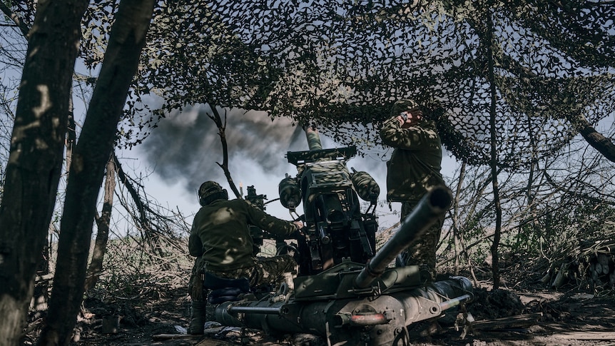 A view of fighters seen from back with weaponry under netting amid trees.