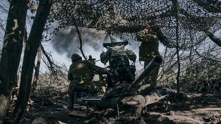 Soldiers tend a stationary cannon underneath camouflage netting.