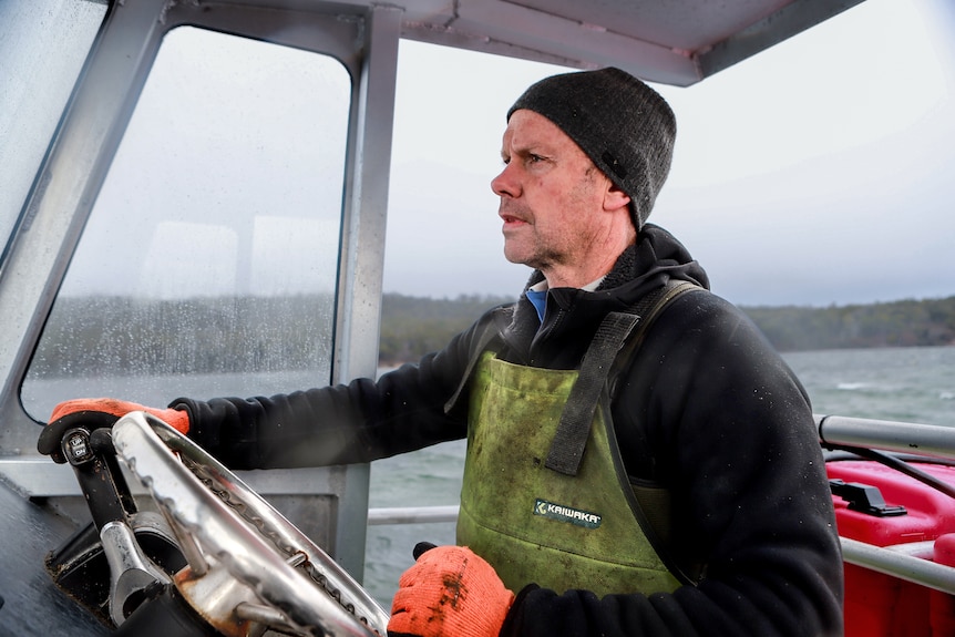 A man wearing a beanie and warm waterproof clothes looks out through a small boat window with hands on a steel steering wheel