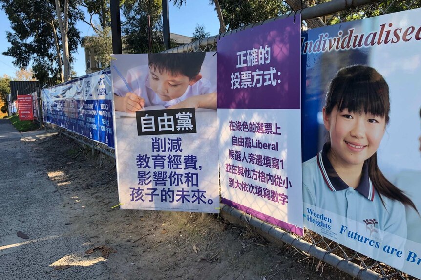 Two rival signs showing mandarin text on a chain fence, one depicts a Chinese child writing with pen and paper.