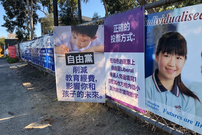 Two rival signs showing mandarin text on a chain fence, one depicts a Chinese child writing with pen and paper.