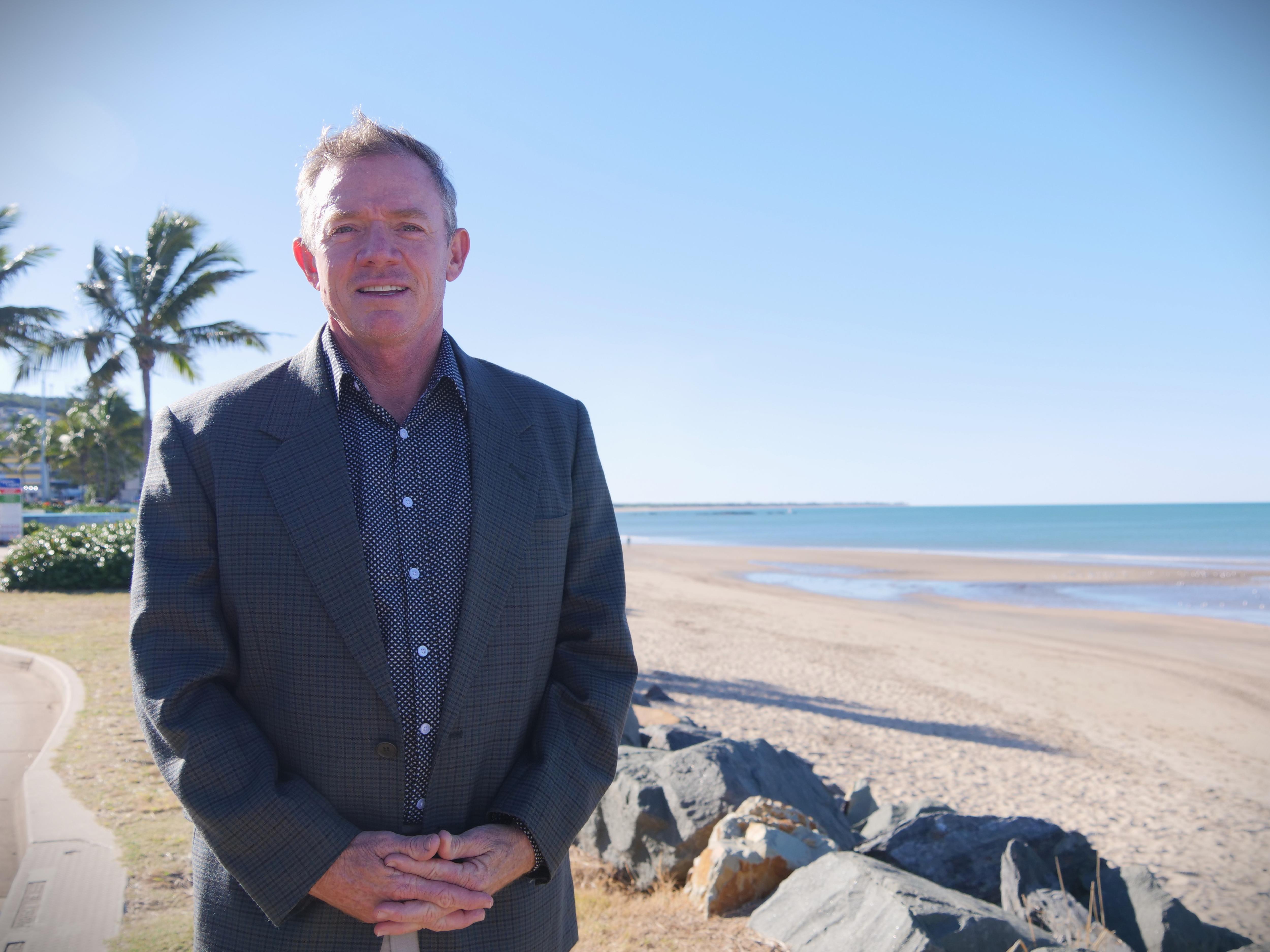 A man in a button up shirt and suit jacket standing in front of a beach