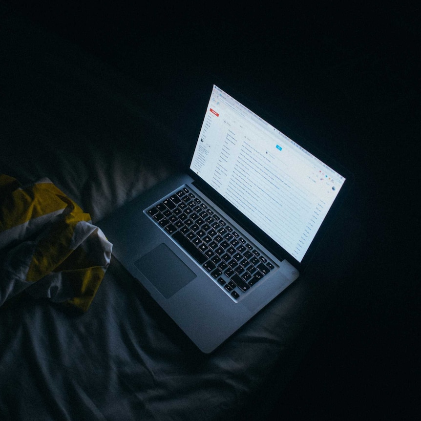 A laptop computer sits open on a bed in a dark room showing an email account on screen.