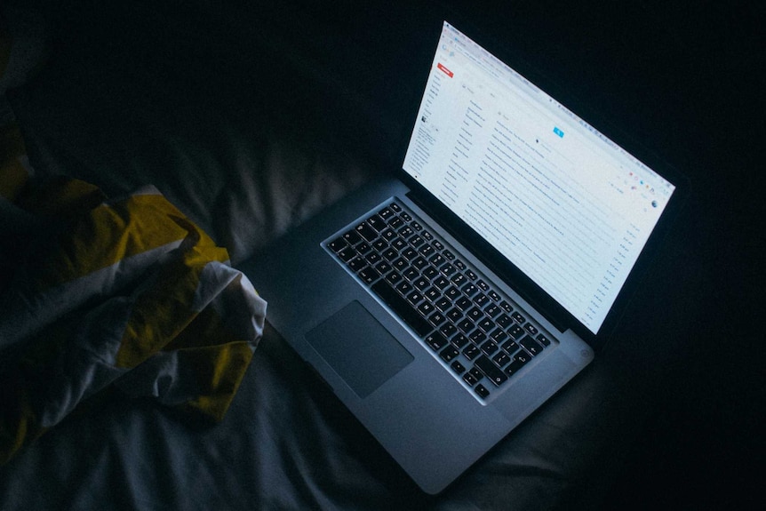 A laptop computer sits open on a bed in a dark room showing an email account on screen.