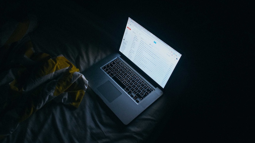 A laptop computer sits open on a bed in a dark room showing an email account on screen.