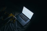 A laptop computer sits open on a bed in a dark room showing an email account on screen.
