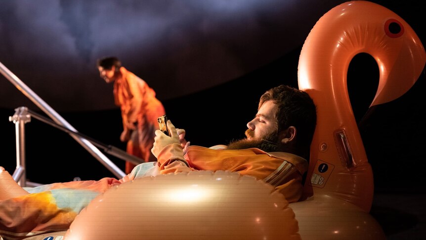 Scott Price, in an orange tracksuit, sits in an inflatable flamingo looking at a smartphone.