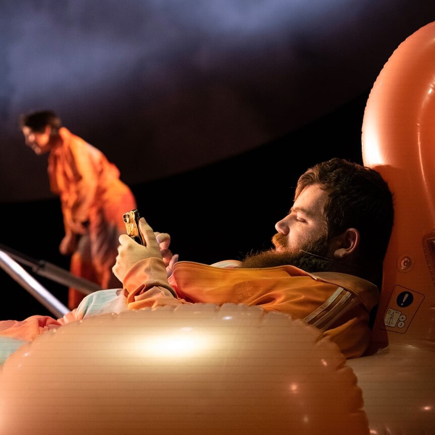 Scott Price, in an orange tracksuit, sits in an inflatable flamingo looking at a smartphone.