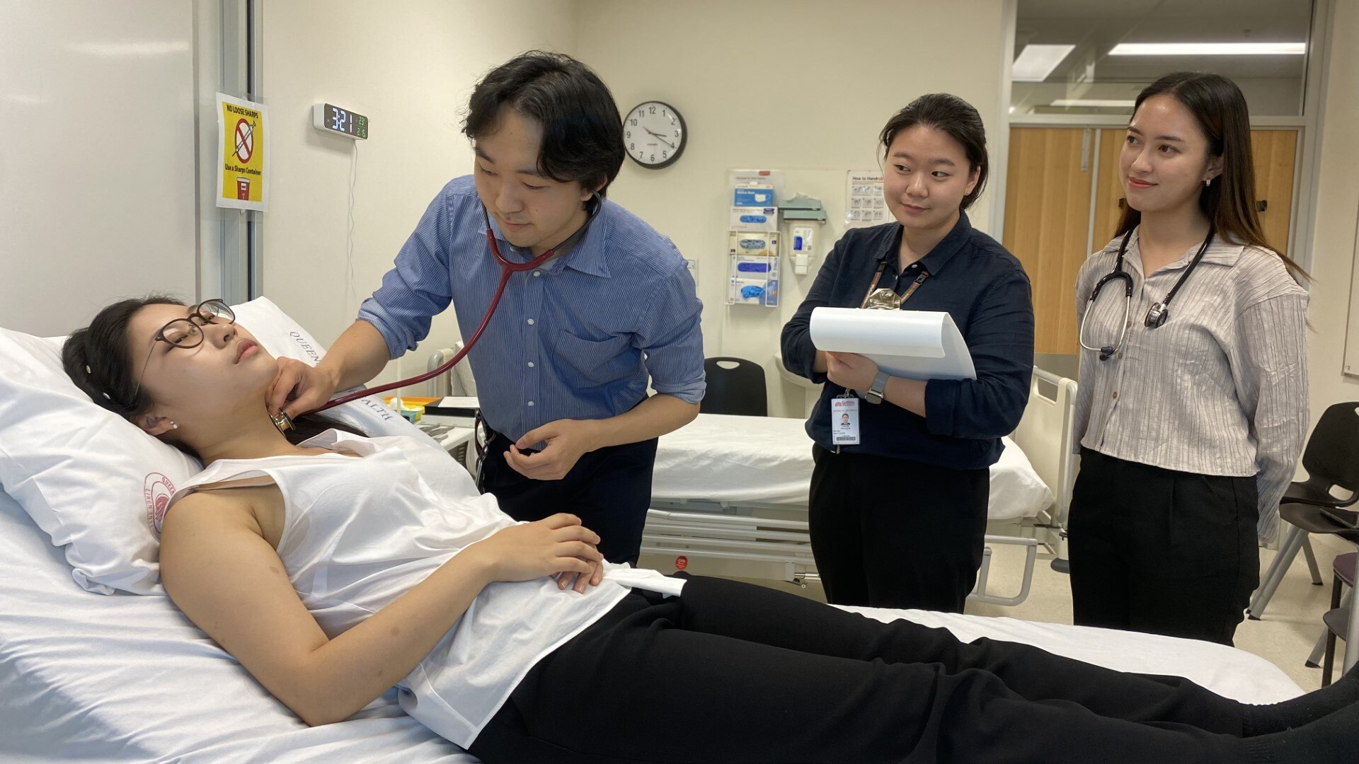 A young man uses a stethoscope to monitor a woman lying on a bed while a group of others watch on.