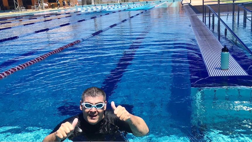 A man wearing goggles and a black vest in a blue swimming pool with thumbs up.