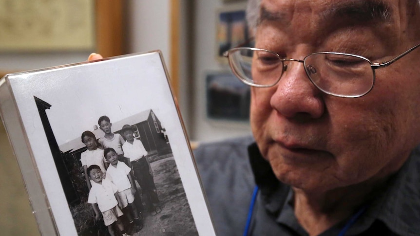 An elderly man holds a black-and-white photo of him as a child with his family