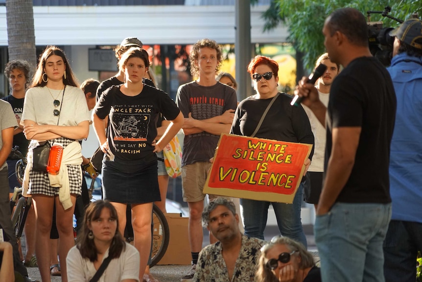 A group of people stand watching a man with a microphone. One person is holding a sign saying WHITE SILENCE IS VIOLENCE