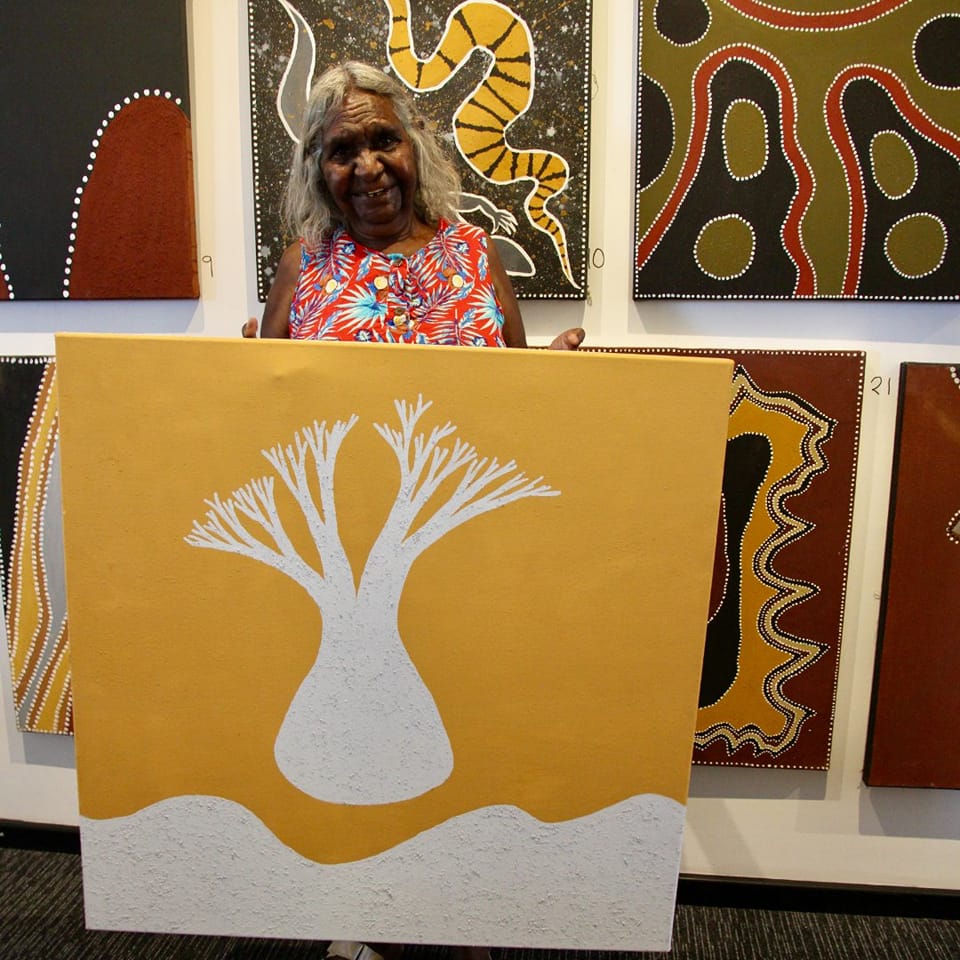 A woman smiles while holding a white and yellow canvas with a tree