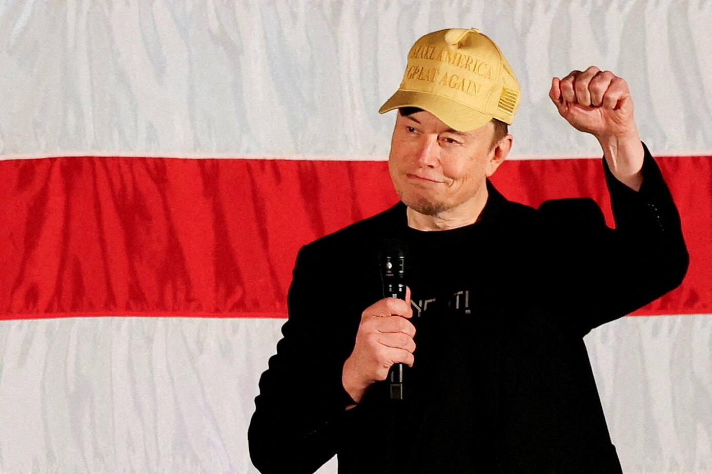 Elon Musk wearing a dark suit blazer and shirt and a gold Make America Great Again cap holding a microphone and raising a fist