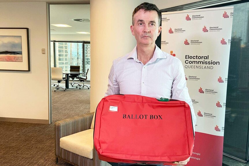 El comisionado del ECQ, Pat Vidgen, sostiene una urna roja en la oficina del ECQ en Brisbane, cabello corto, camisa clara, serio.