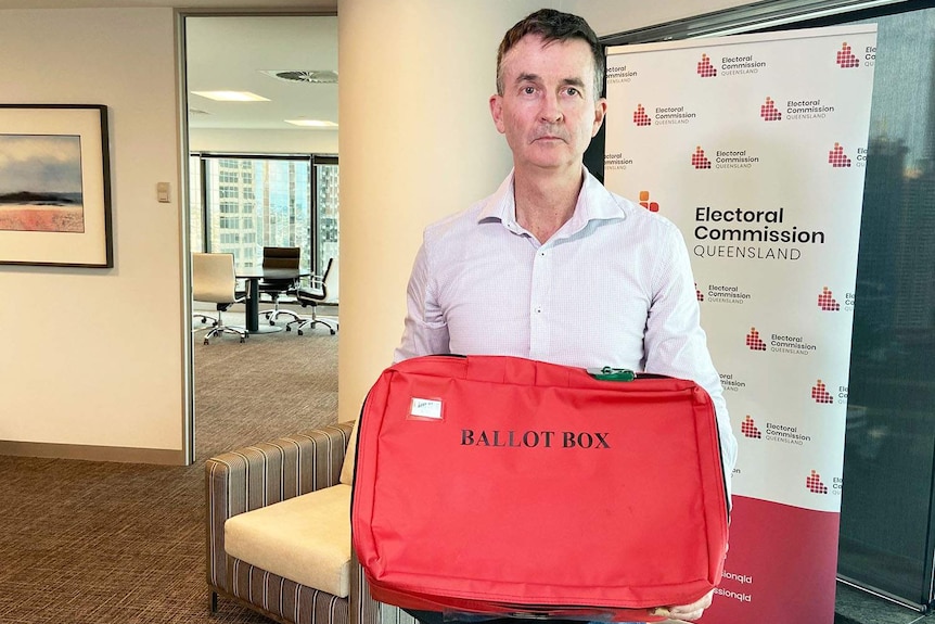ECQ Commissioner Pat Vidgen holds a red ballot box case in the ECQ office in Brisbane, cropped hair, light shirt, serious.
