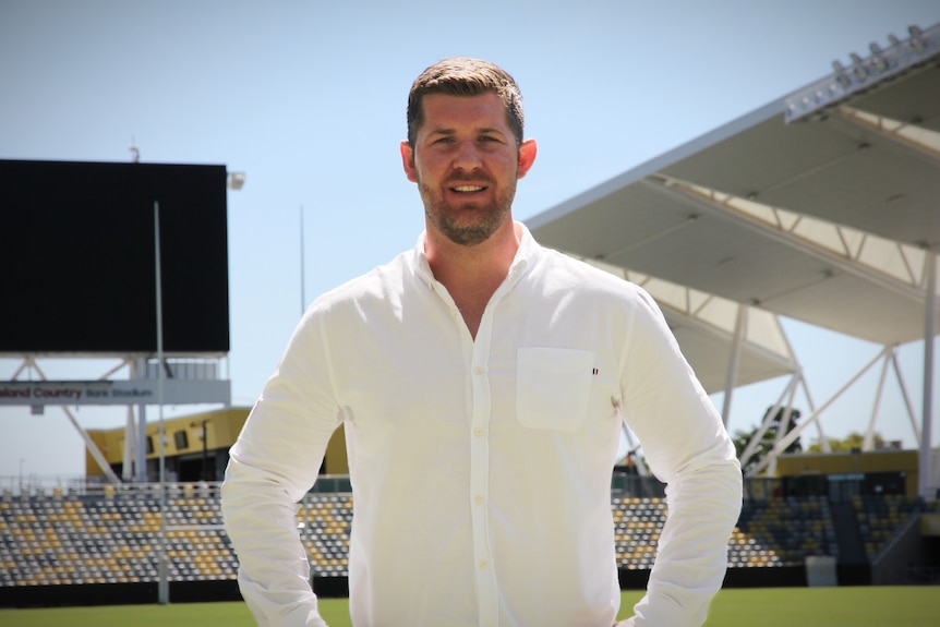 Un homme souriant vêtu d'une chemise blanche se tient sur un champ à l'intérieur d'un stade vide, les mains sur les hanches. 