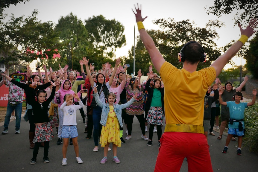 A group of children and adults wearing headphones dance with their hands in the air