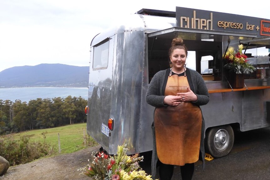 A woman stands in front of a coffee van, the sea and mountains in background.
