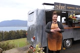 A woman stands in front of a coffee van, the sea and mountains in background.