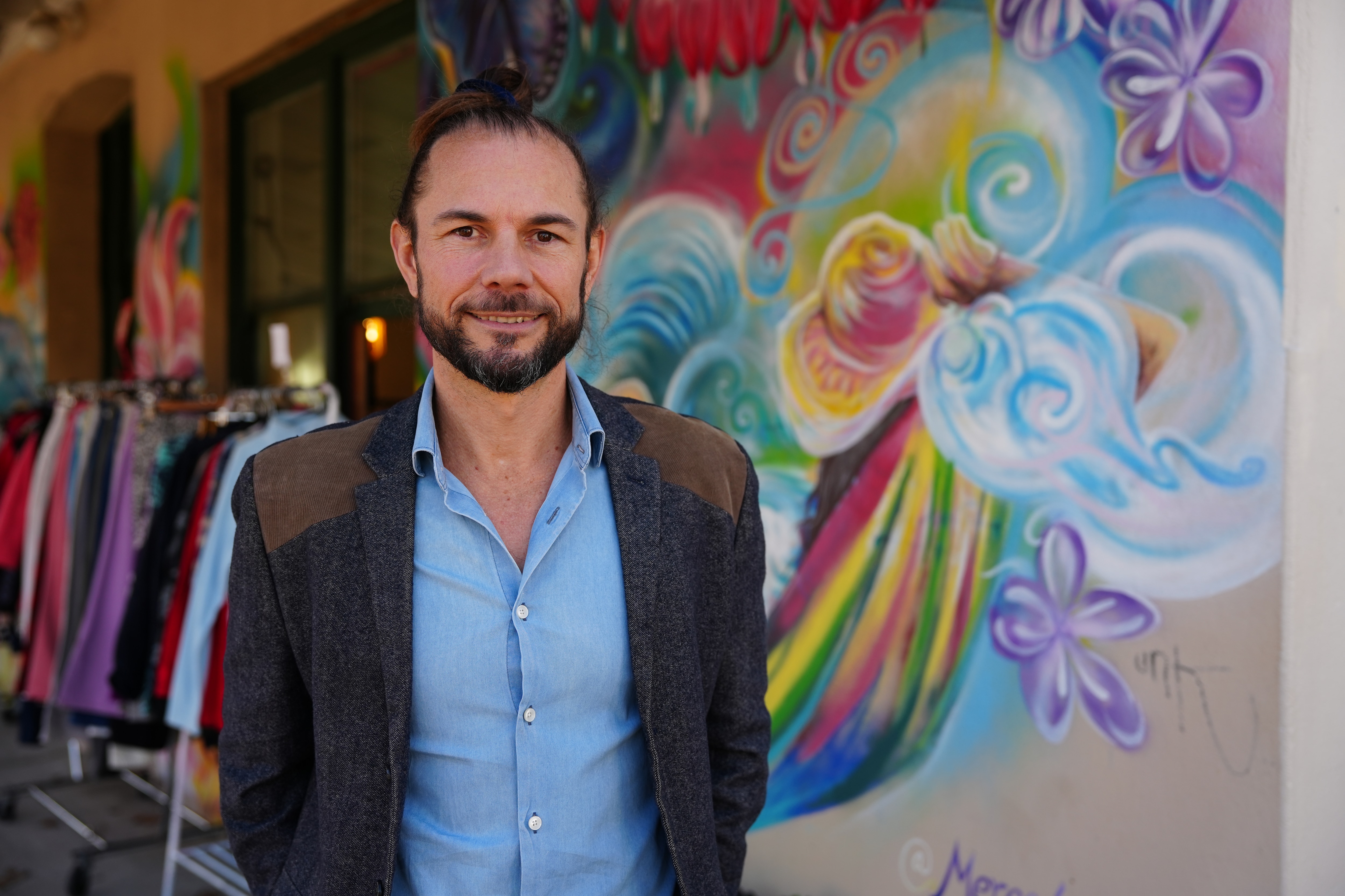 Man in a blue business shirt and grey jacket standing near a colourful wall mural.