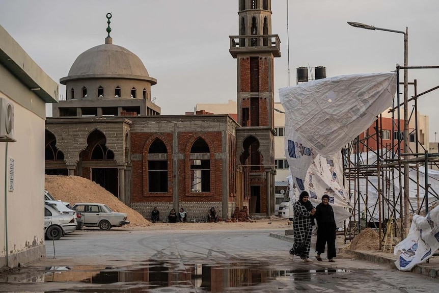 Two people walk on a street near some scaffoling and a mosque.