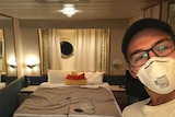 A man wearing a hat, glasses and a face mask. He is in a small cabin room with a port window and a bed.