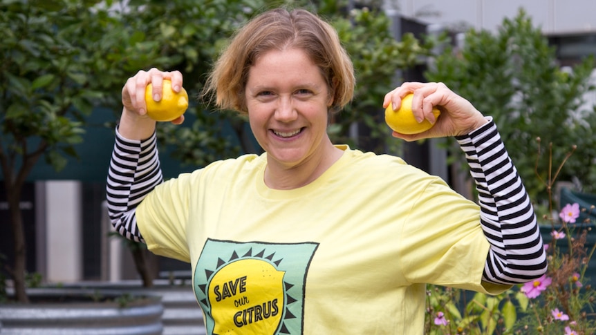 A woman, wearing a yellow t-shirt saying 'Save Our Citrus', makes a strong-arm pose while holding lemons.