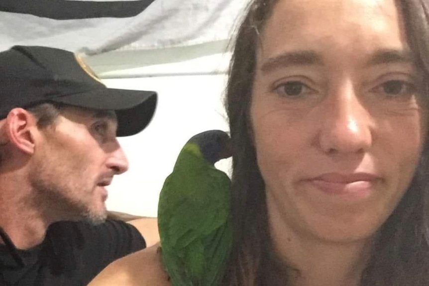 A man and woman pose for a selfie with a small green parrot on the woman's shoulder.