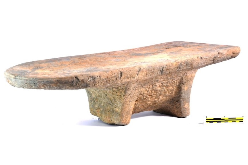 A large ancient stone grinding tool that looks like a surfboard-shaped table with two thick legs.