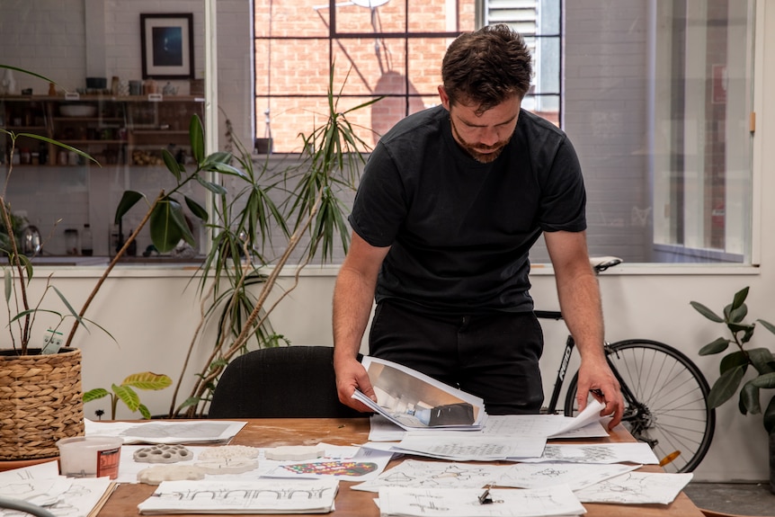 Alex Goad leans over a wooden table, and sorts through a litany of with A4 documents and sketches.