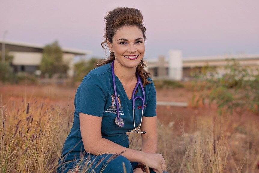 A female doctor dressed in scrubs sits on grass.