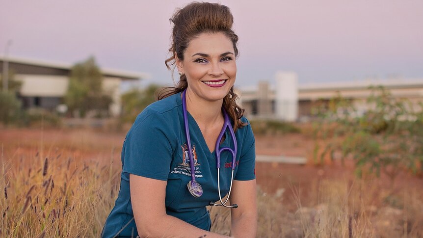A female doctor dressed in scrubs sits on grass.