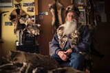 A man with a big grey beard sits in a room surrounded by feral cat skins looking blankly at the camera