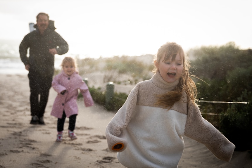 Two young girls run ahead of their dad laughing and smiling on a beach path with the sun's glare behind them.