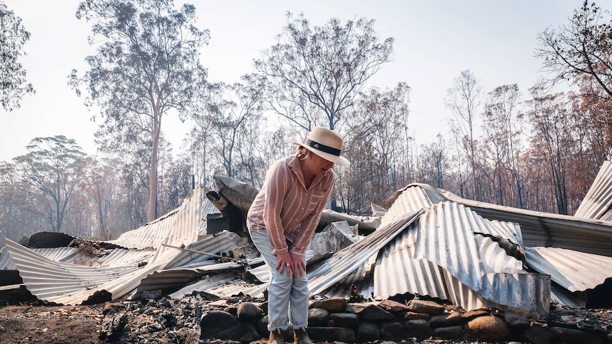 Melinda Plesman stands among twisted corrugated iron and ashes, with burnt trees and smoky haze in the background.