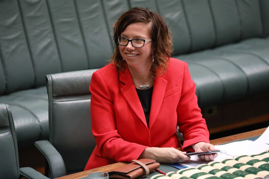 Amanda Rishworth, wearing a red blazer, smiles sits in her seat. She is holding her mobile