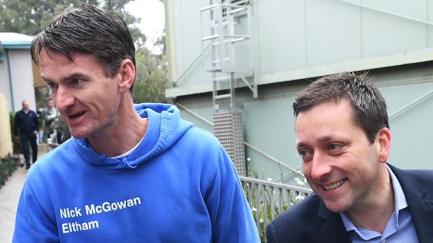Nick McGowan, in a blue hoodie reading 'Nick McGowan Eltham' next to a smiling Matthew Guy in a suit, both reaching out hands.