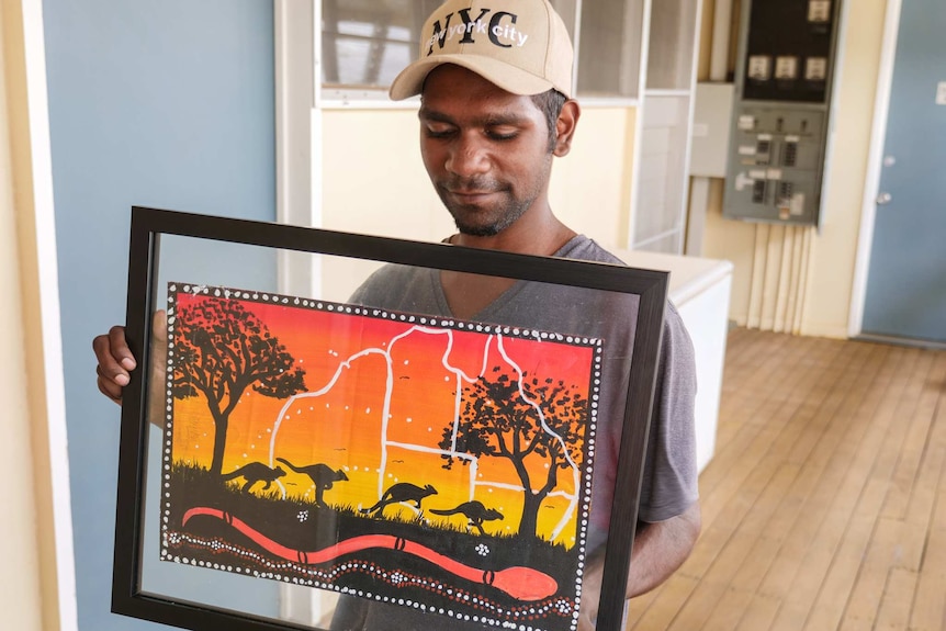 A young indigenous man holds a framed painting of blacks, reds, oranges, and yellows, which shows a kangaroo in the outback.