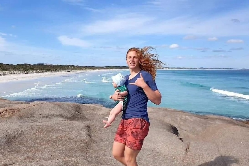 A man with long red hair stands on rocks near the ocean, while holding a toddler.
