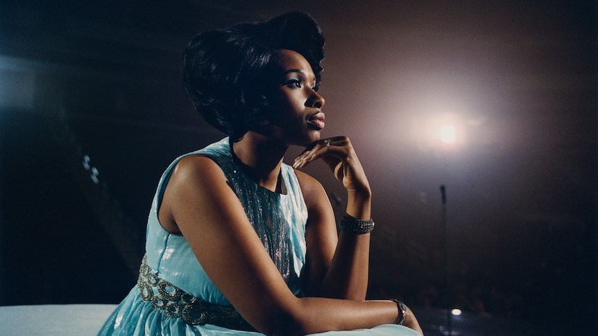 Jennifer Hudson sits in a pale blue 50s style dress, resting her head in her hands and looking pensively into the distance.