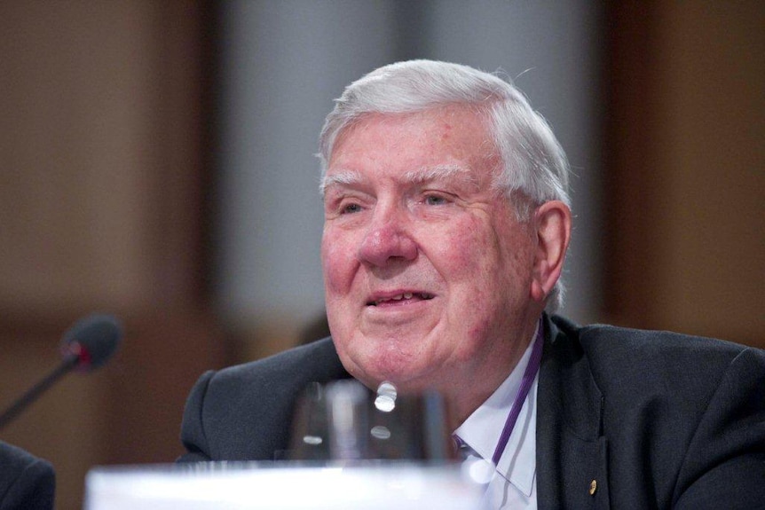 Headshot of elderly man wearing suit, spekaing into microphone