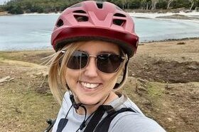 A smiling blonde woman with a helmet on, riding her bicycle near a lake.