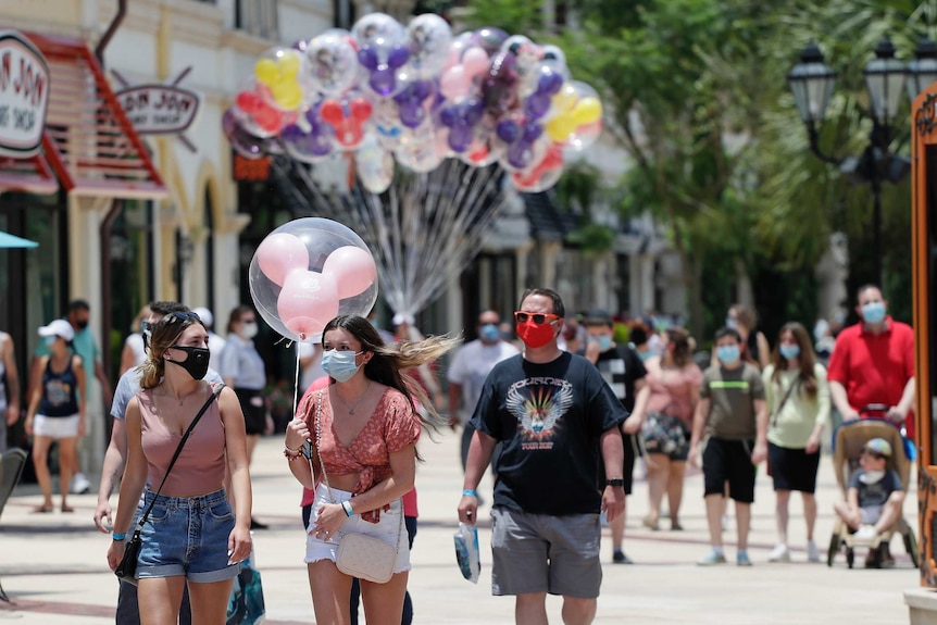 A woman holding a balloon wears a face mask as she walks through a busy pedestrian area in a theme park