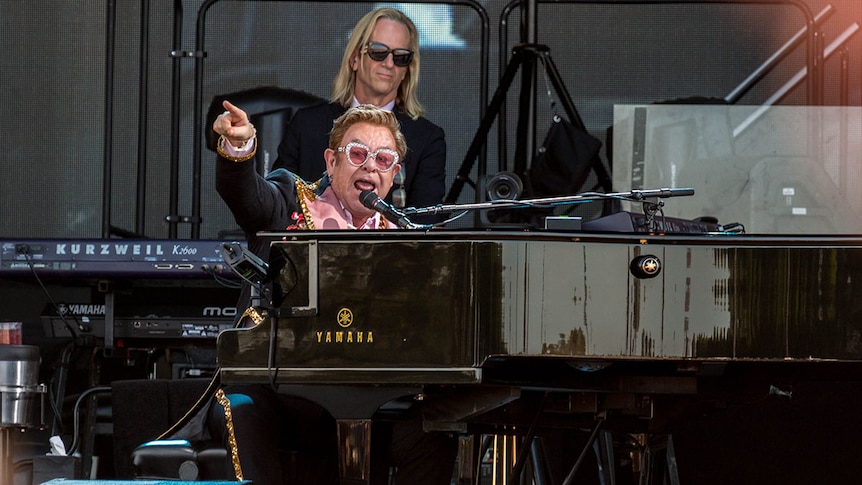 Performer Sir Elton John sits at his piano on a stage and points to the crowd.