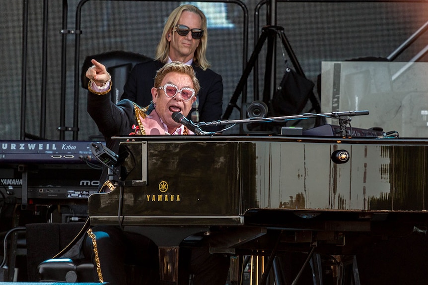 Performer Sir Elton John sits at his piano on a stage and points to the crowd.